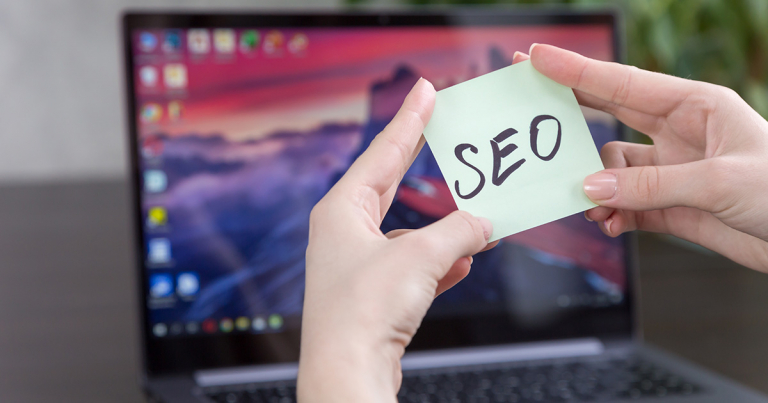 Le mani di una donna tengono un post it con la scritta SEO davanti ad un PC portatile